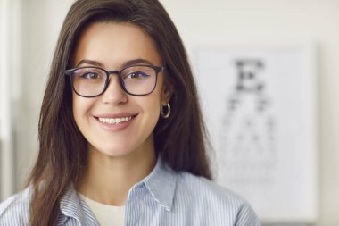 Çok güzel gülümseyen genç bir kadının portresi kameraya bakıyor ve göz doktorunda gözlük takıyor ya da arka planda göz çizelgesi olan optik bir dükkan. Sağlık ve Tıp Konsepti.