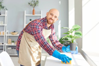 Ev temizliği sırasında mobilyaların tozunu alan gülümseyen kel bir adamın olumlu portresi. Neşeli tavırlar ev işlerine ve ev işlerine olan bağlılığını yansıtır. Temiz ve düzenli bir ev ortamını garantiler..