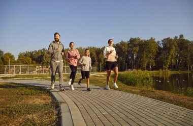 Spor kıyafetlerinde birlikte koşan mutlu aktif aile, açık havada egzersiz yapmanın keyfini çıkarın. Ebeveynler ve çocuklar hafta sonu vakit geçirirler, sağlıklı şehir hayatı, dost canlısı alışkanlıklar.