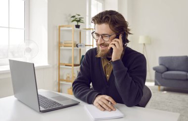 Neşeli iş adamı cep telefonundaki hoş ve başarılı iş konuşmalarından memnun. Telefonda konuşan, gülümseyen sakallı adam masada dizüstü bilgisayar ve not defterinin önünde oturuyor..