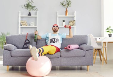 Spor kıyafetleri içinde, elinde bira şişesi ve spor sonrası elinde dambılla evdeki koltukta oturan komik bir genç adam. Mutlu gülümseyen sporcu adam kanepede alkolle dinleniyor.