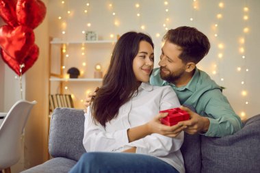 Mutlu çift hediye kutusunu bir arada tutarak birbirlerine bakıyorlar. Karı koca kalp şeklinde balonların arka planında kanepede oturup birbirlerine şefkatle bakıyorlar.