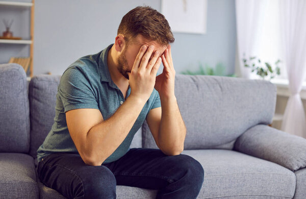 Emaciated, disappointed man passing through relationship problems holds his head with hands and suffering from headache alone on the sofa. Stress, relationship problems, conflicts and divorce concept
