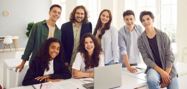 Sınıfta öğretmenle birlikte mutlu lise öğrencilerinin toplu portresi. Neşeli genç oğlanlar ve kızlar ve masada dizüstü bilgisayarlı genç bir öğretmen kameraya bakıyor ve gülümsüyor.