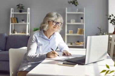 Kıdemli gülümseyen kadın çalışma masasında oturmuş dizüstü bilgisayarla çalışıyor ve planlama bütçesini not alıyor. Yetişkinler için e-öğrenme kavramı ya da emeklilik için serbest meslek anlayışı.