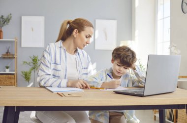 Kendine güvenen genç bir anne, küçük oğluyla birlikte evdeki masada oturuyor ve ona ödevlerinde yardım ediyor. Uzaktan ders sırasında bilgisayarda ders çalışan bir çocuk. Eğitim ve bilgi kavramı.
