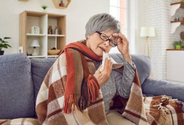 Sick senior woman with runny nose wrapped in a blanket sitting on sofa at home. Elderly person with seasonal flu sneezing in napkin, feel unhealthy with influenza and touching her forehead with hand.