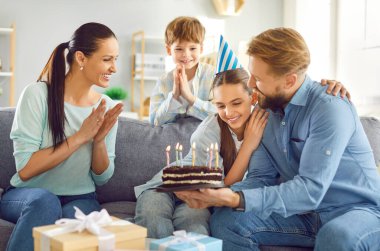 Mutlu gülümseyen, parti şapkalı ergen kız çocuğu evde anne, baba ve erkek kardeşle birlikte mumlarla pastaya bakarak dilek tutuyor. Mutlu aile ve bayram kutlamaları.