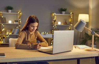 Beyaz kız öğrenci bir dizüstü bilgisayarla masada oturuyor, ödev yapıyor ve bir deftere yazıyor. Akşamları evde eğitim görüyor, öğrenim görüyor ve okul ödevlerini internet üzerinden tamamlıyor..