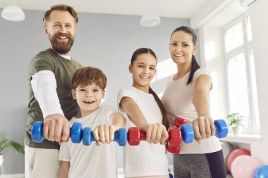 Spor salonundaki spor eğitimi sırasında elinde dambıl tutan çocuklu bir ailenin portresi. Birlikte gülümsüyor ve spor faaliyetlerinden zevk alıyorlar, aile olarak sağlıklı bir yaşam tarzını destekliyorlar..