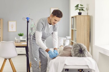Genç, dost canlısı hemşire, klinikte serumdan serum verilmesi ve vitamin terapisi gören yaşlı hasta kadına yardım ediyor. Erkek doktor, yaşlı hastasına danışmanlık yapıyor. Hastanede kanepede yatıyor..