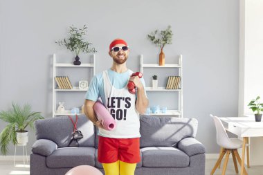 Spor kıyafetleri giyen, yoga minderi tutan, elinde dambıllar olan ve oturma odasında kameraya bakan komik bir gencin portresi. Spor, spor ve ev eğitimi konsepti.