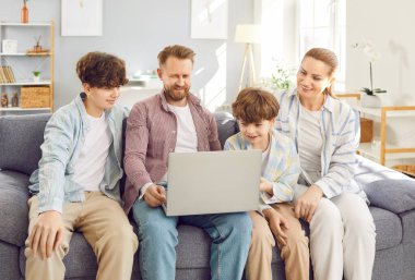 Mutlu bir aile, genç ve küçük oğulları olan çocuklar kanepeye oturmuş birlikte yeni bir dizüstü bilgisayar kullanarak eğleniyorlar, gülümseyen ebeveynler haftasonunun tadını çıkarmak için kanepede dinleniyorlar, evde bilgisayar başında video izliyorlar.