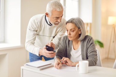 Yaşlı kadın, yaşlı bir adamın elinde tuttuğu akıllı telefon ekranından ayrıntıları dikkatlice kopyalıyor. Modern teknolojiler ve aile ortaklığı ile yaşlı insanların etkileşimi kavramı.