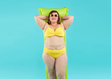 Mutlu komik artı güneş gözlüklü kadın ve yazlık sarı bikinili stüdyo mavisi arka planda izole edilmiş şişme yatakta yatan kadın. Şişman kız plajda yüzmeye gidiyor. Tatil kavramı.