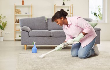 Mutlu orta yaşlı Afro-Amerikan kadın diz temizleme halısı ve modern kablosuz elektrikli süpürgeyle mobilya, profesyonel temizlik işçisi, hizmetçi, ev işi yapan hademe, ev hanımı.