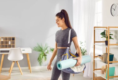 Gülümseyen atletik kadın evde spor çantasıyla dolu bir spor çantası tutuyor. Spor salonunda antrenman yapmaya hazır. Kendini spora, yogaya ve aktif bir yaşam tarzına adamış..