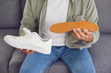 Evdeki oturma odasında kanepede oturan ve elinde beyaz spor ayakkabılarla ortopedik ayakkabı tabanı tutan gündelik giysiler giyen bir erkek. Ayaklar rahat ve sağlık hizmeti konsepti.