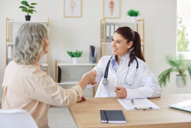 Klinikteki tıbbi muayeneden sonra masada oturan yaşlı bir kadınla tokalaşan güler yüzlü kadın doktor terapisti. İlaç ve sağlık hizmetleri kavramı.