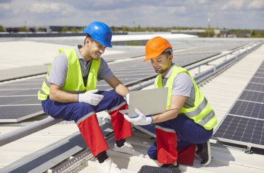 Güneş panellerindeki teknisyenler çatıda bir dizüstü bilgisayar kullanarak montaj ya da onarım işiyle meşguller. Fotovoltaik sistemlerin teknolojisi, sürdürülebilir güç ve enerji çözümlerine katkıda bulunuyor.