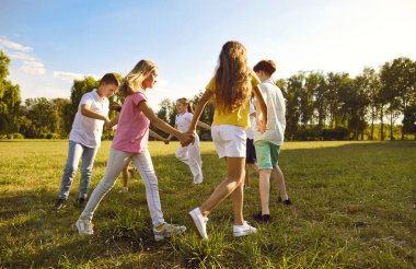Dışarıda parkta oynayan bir grup çocuk bir çember oluşturup el ele tutuşuyorlar. Güneşli bir yaz gününde gündelik kıyafetlerle oyun oynayan ve dışarıda eğlenen mutlu çocuklar..