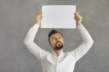 Elinde boş bir kağıt pankart tutan şüpheyle dolu bir gencin stüdyo portresi. Ciddi bir adam gri bir arka planda tek başına duran boş, beyaz bir panoya bakıp düşünüyor.