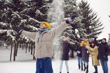 Küçük şeylerden zevk al. Neşeli ve kaygısız genç adam, arkadaşlarının ona kartopu attığı arka planda kollarını açmış duruyor. Arkadaşlar birlikte vakit geçirir ve karlı ormanda eğlenirler..