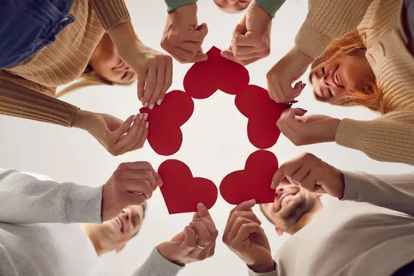 stock image Bottom view of a happy smiling group of people holding red hearts in hands celebrating valentines day together. Young friends putting their arms in a circle. Unity, love and friendship concept.