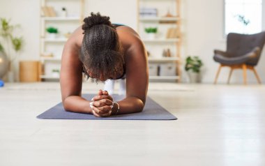 Evdeki oturma odasının zemininde yoga minderinin üzerine uzanmış, tahta sporu yapan genç, şişman, Afro-Amerikan bir kadının portresi. Fitness, fit, spor ve ev eğitimi konsepti.