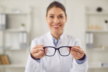 Gülümseyen bir göz doktorunun portresi, göz bakımı konusunda uzman bir doktor, klinikte gözlük tutuyor. Optalmoloji alanında profesyonel sağlık hizmetleri. Göz bezi reçetesi.