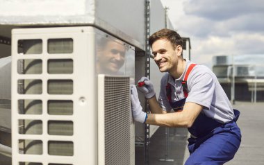 Genç profesyonel teknisyen üniforma giyiyor ve çatıda oturan tornavidayı kullanarak klima kuruyor ve tamir ediyor. Gülümseyen erkek tamirci havalandırma sistemini tamir ediyor..