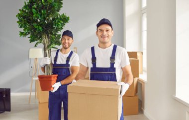 Gülümseyen genç adamların portresi üniformalı işçilerin elinde bir karton kutu ve boş bir dairede duran kameraya bakan yeşil bitki. Taşıma, taşıma ve taşıma hizmeti kavramı.