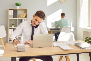 Neşeli, gülümseyen, beyaz gömlekli ve kravatlı bir iş adamı iş yerinde oturmuş cep telefonuyla konuşuyor ve notlar alıyor. Modern ofiste çalışan iki genç erkek şirket çalışanı..