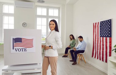 Oylayın. Amerikan bayrağıyla süslenmiş oy verme kabininin yanında duran sorumlu kadın seçmenin portresi. Genç bir kadın sandık merkezinde poz veriyor ve kameraya gülümsüyor. Amerika 'da seçim kavramı.