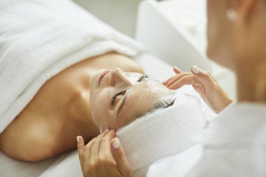 Güzellik uzmanı ya da kozmetikçi, güzellik salonundaki kırışıklık önleyici kozmetik işlemleri için genç kadın yüzüne nemlendirici çarşaf maskesi uyguluyor. Kozmetoloji ve profesyonel cilt bakımı kavramı.