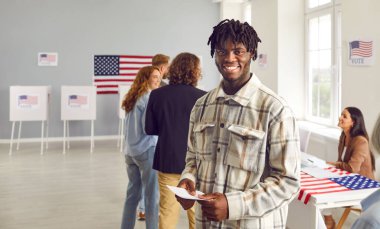 Genç mutlu Amerikalı seçmen oy merkezinde kuyrukta bekliyor ve seçim günü oy kullanma merkezindeki kameraya neşeli bir şekilde bakıyor. Seçim merkezinde oy pusulası tutan adam.