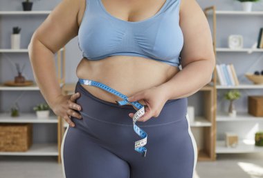 Şişman, spor giysili bir kadının yakın plan çekilmiş fotoğrafı. Evdeki oturma odasında duran ölçü bandıyla belini ölçün. Obezite, ağırlık kontrolü, diyet ve zayıflatma kavramı.