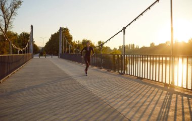 Gün batımında şehir parkında köprüden koşan zayıf, genç bir kadın. Spor giyimli atlet kız dışarıda tek başına koşuyor. Sağlıklı yaşam tarzı, spor çalışması ve doğa konseptinde uygunluk..