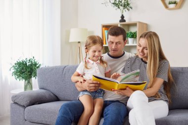 Genç, neşeli bir aile birlikte kanepede oturup kızlarına kitap okuyor. Baba dizlerinin üzerine çöküp çocuğu kucağına alıyor ve anne kitaptaki resimleri gösteriyor. Öğrenme, hikaye anlatma ve çocuk gelişimi.