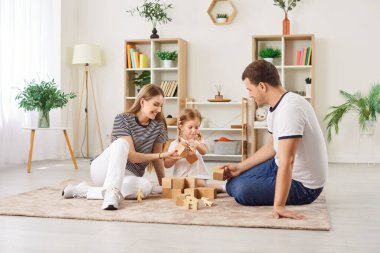 Neşeli ebeveynler kızlarıyla oturma odasında tahta blok oyunu oynuyorlar, anaokulu çocuklarına oyuncak kulesi yapmaları için yardım ediyorlar. Evde aile aktiviteleri. Eğlenmek, yaratıcılığı öğrenmek..