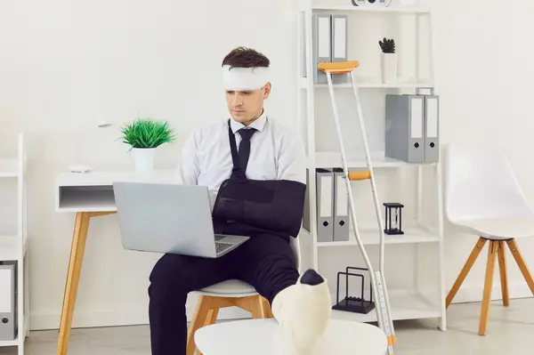 Stock image Handicapped young business man working on a laptop at office or at home sitting on chair with bandaged head and a crutch. Male injured employee with a broken arm and leg on workplace.