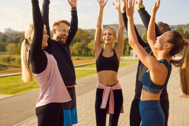 Bir grup mutlu sporcu bir parkta spor ve spor yapıyor. Elleri havada bir çember halinde duran takım, antrenman sırasında takım çalışmasını ve beraberliği gösteriyor..