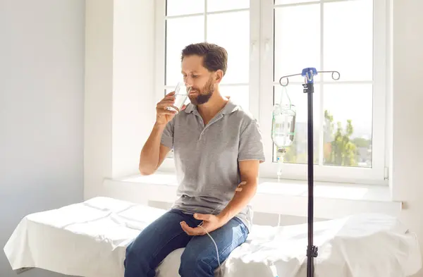 stock image Young man sitting on the couch at doctors office in clinic while receiving IV drip infusion and vitamin therapy in his blood. Male person receiving injection therapy and drinking a glass of water.