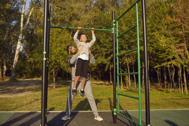 Genç bir baba ya da erkek koç, çocuk bir çocuğa dışarıdaki yaz parkında kendini yukarı çekmesine yardım ediyor. Sportif aile doğada spor egzersizleri yapıyor. Sağlıklı yaşam tarzı kavramı.