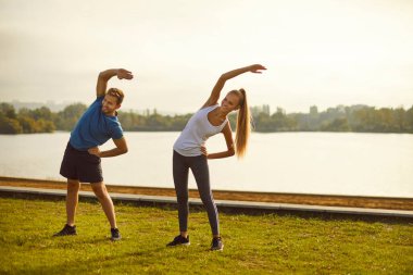 Genç, mutlu, gülümseyen bir çift doğada spor germe egzersizi yapıyor. Spor giyinen erkek ve kadın sporcu parkın dışında spor yapıyorlar. Sağlıklı yaşam tarzı ve egzersiz konsepti.