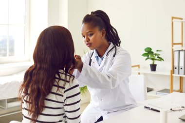 Tıbbi laboratuvar önlüklü siyahi kadın doktor hastane muayenesi sırasında bir hastayı muayene ediyor. Hemşire hastanın endişelerini dinler ve etkili sağlık hizmetleri için hayati önem taşıyan açık diyalogları teşvik eder..