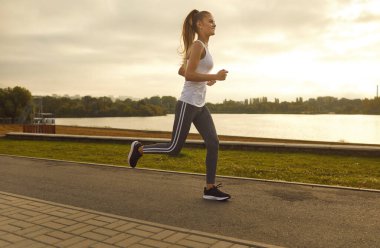 Neşeli, zayıf kız sporcu sabah koşusu sırasında spor yapıyor. Spor tişörtlü, taytlı ve spor ayakkabılı, parkta koşan güzel beyaz bir kadın. Vücut bakımı kavramı.