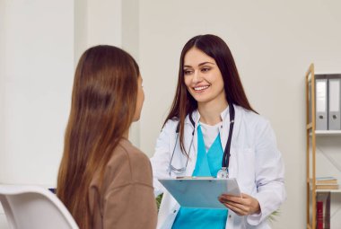 Steteskop takan gülümseyen doktorun portresi randevu ile birlikte rapor tutuyor ve kız hastaya danışmanlık yapıyor. Genç çocuk klinikte tıbbi muayene sırasında doktoru dinliyor.