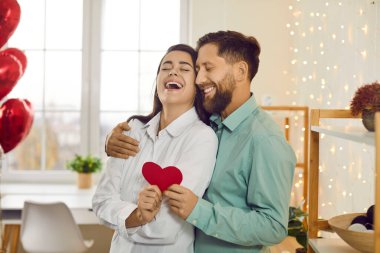 Genç, mutlu, gülümseyen bir adam ve kadının portresi. Ellerinde kalp, kapalı gözlerle sarılmak ve evde kahkaha atmak. Aşık mutlu bir çift. Dikkat, şefkat ve sevgililer günü konsepti.