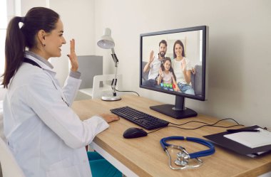 Masasında steteskoplu aile doktoru gülümsüyor ve masaüstü bilgisayar ekranına selam veriyor teletıp danışmanlığında ebeveynleri ve çocukları çağdaş uygun web kamerası uygulaması üzerinden selamlıyor.